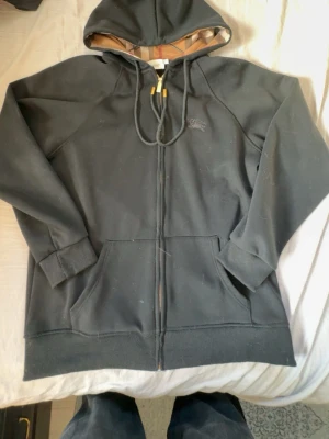 Svart hoodie från Burberry - Säljer en svart hoodie från Burberry, bara testad!! Passar ej mig. Den har dragkedja och guldiga detaljer på snörningen. Hoodien har klassisk huva med Burberrys rutiga mönster på insidan, två stora fickor framtill och ribbade muddar. Priset kan absolut diskuteras 🤝