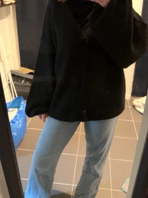 Svart kofta från Nelly - Mysig  kofta från Nelly med v-ringning och knäppning framtill. Koftan har en lös passform och långa ärmar, perfekt att slänga över en t-shirt eller topp. Materialet är mjukt och fluffigt för extra komfort. Helt ny