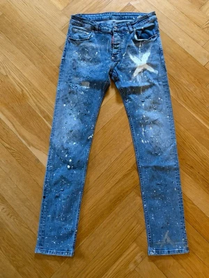 Blå dsq jeans  - Unika blå jeans med smal passform, dekorerade med vita och ljusa färgstänk samt en stor vit stjärna på vänster lår. Klassisk femficksmodell och knappgylf. Perfekta för dig som gillar en kreativ och personlig stil.