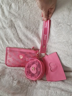 Juicy Couture rosa plånboksset - Säljer ett rosa plånboksset från Juicy Couture med tre delar: en genomskinlig plånbok med logga, en rund myntbörs med hjärtdetalj och en korthållare med präglat hjärta. Alla delar sitter ihop med en bred handledsrem med Juicy-logga. Perfekt för dig som gillar rosa accessoarer!