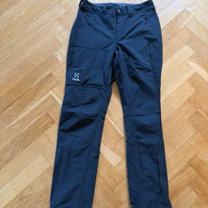 Svarta byxor från Haglöfs - Snygga svarta byxor från Haglöfs med raka ben och praktiska fickor, inklusive en benficka med logga. Tillverkade i slitstarkt syntetmaterial, perfekta för dig som gillar outdoor och funktionell stil.