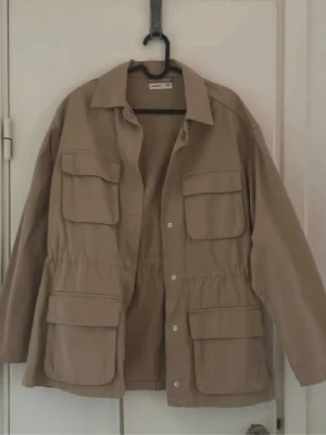 Beige overshirt från Perfect Jeans - Stilren beige overshirt från Perfect Jeans med fyra stora fickor framtill och knappar hela vägen. Overshirten har en avslappnad passform, klassisk krage och är gjord i ett mjukt bomullsliknande material. Perfekt att slänga över en hoodie eller t-shirt.