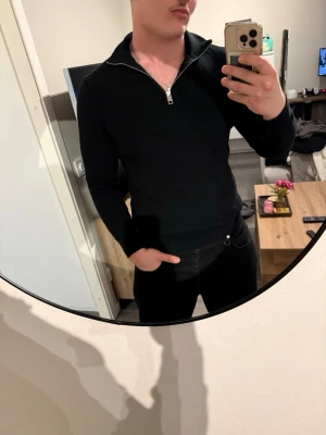 Svart stickad tröja med dragkedja H&M - Svart stickad tröja från H&M med halv dragkedja och hög krage. Tröjan har lång ärm och ribbstickad struktur, perfekt för lager-på-lager. Enkel och stilren design som passar till det mesta.