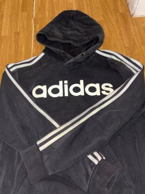 Svart Adidas hoodie  - Svart hoodie från Adidas med stor vit logga på bröstet och klassiska vita ränder längs ärmarna. Tröjan har huva med dragsko och en magficka. Perfekt för en sportig och avslappnad stil.