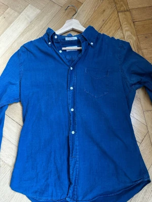 Blå oxfordskjorta från Gant Rugger - Snygg blå oxfordskjorta från Gant Rugger med klassisk button-down krage och bröstficka. Skjortan är tillverkad i bomull och har vita knappar framtill. Perfekt för en stilren och avslappnad look.