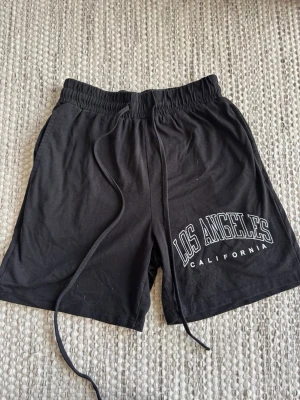 Svarta shorts med Los Angeles-tryck - Svarta shorts med resårmidja och snörning samt trycket 'Los Angeles California' i vitt på ena benet. Sköna och avslappnade, perfekta för sommaren. Tillverkade i mjuk bomull för maximal komfort. Prisförslag tas emot 💗