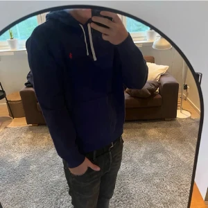 Mörkblå hoodie från Ralph Lauren - Säljer en mörkblå hoodie från Ralph Lauren med vit dragsko i huvan och den klassiska röda loggan broderad på bröstet. Hoodien har en avslappnad passform och är perfekt för en chill look. Materialet känns mjukt och bekvämt.