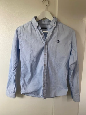 Ljusblå skjorta från U.S. Polo Assn - Klassisk ljusblå skjorta från U.S. Polo Assn med broderad logga på bröstet. Skjortan har knappar framtill och lång ärm. Perfekt för en stilren och avslappnad look.