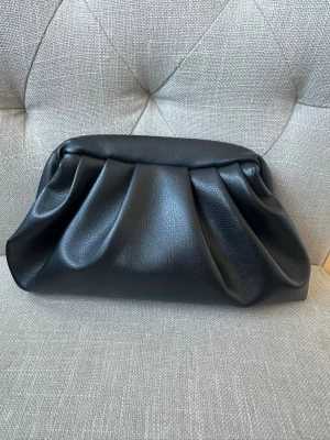 Svart clutchväska i konstläder - Stilren svart clutchväska med mjuk, veckad design i skinnimitation. Väskan har en rymlig öppning med tygfoder och stängs med metallspänne. Perfekt accessoar för dig som gillar minimalistisk och trendig look. Väskan är handgjord 