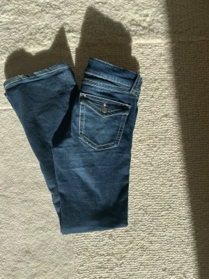 Mörkblå bootcut jeans med kontrastsömmar - Snygga mörkblå bootcut jeans med vita kontrastsömmar och klassiska fem fickor. Jeansen har en knappstängning och detaljerad bakficka med lock och knapp. Perfekta för en avslappnad och trendig look.