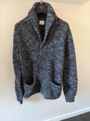 Gråmelerad kofta från Polo Ralph Lauren - Mysig gråmelerad kofta med grovstickad känsla från Polo Ralph Lauren. Koftan har stora fickor framtill, knäppning med fyra knappar och en hög krage. Perfekt för lager-på-lager och chill dagar.