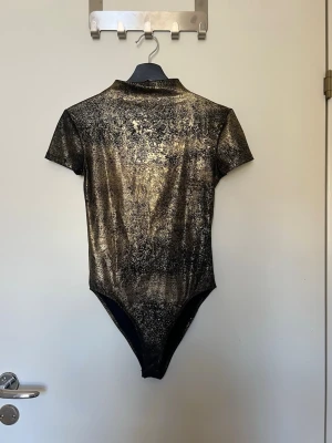 Svart och guld body från Zara - Snygg body från Zara i svart med skimrande guldmönster. Kortärmad modell med hög krage och stretchigt material som sitter tight mot kroppen. Perfekt för dig som vill sticka ut med en glittrig look. Materialet känns mjukt och följsamt.