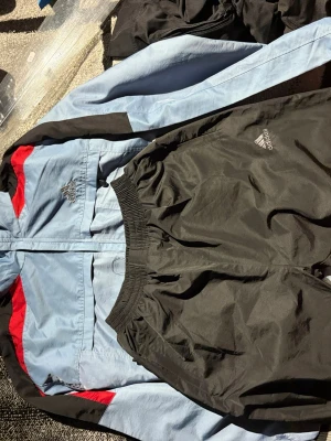 Adidas träningsset blå/svart/röd M-L - Snyggt träningsset från Adidas med en ljusblå jacka med svarta och röda detaljer samt matchande svarta byxor. Jackan har dragkedja, ståkrage och två fickor. Adidas-logga på både jacka och byxor. Materialet är lätt och passar perfekt till träning.