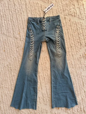 Blå snörade jeans från I.AM.GIA - Unika blå jeans från I.AM.GIA med vita snörningsdetaljer längs benen och framtill. Jeansen har utsvängda ben och är lågmidjade. Mått: Midja (lågmidjade!) 38cm Innerbenslängd (82/83cm lite osäker eftersom dom är klippta så ojämnt där nere)