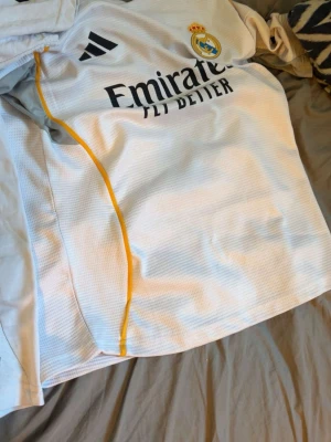 Real Madrid träningströja Adidas M - Vit Real Madrid träningströja från Adidas i slim fit-modell. Tröjan har svart Adidas-logga, klubbmärke och sponsortryck framtill samt gula detaljer längs sidan. Tillverkad i ett ventilerande, tekniskt material som passar perfekt för träning.
