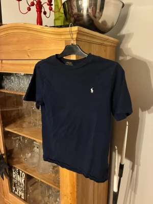 Mörkblå t-shirt från Polo Ralph Lauren - Snygg mörkblå t-shirt från Polo Ralph Lauren med klassisk passform och korta ärmar. T-shirten har en liten vit broderad logga på bröstet och är gjord i mjuk bomull. Perfekt till jeans eller shorts för en clean look. Använd ett fåtal gånger och har storleken L