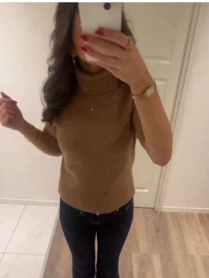 Beige stickad polotröja med kort ärm - Snygg beige stickad polotröja med korta ärmar och ribbade kanter. Tröjan har en hög krage och är perfekt att styla med jeans eller kjol. Materialet ser mjukt och bekvämt ut, och färgen är lätt att matcha med andra plagg.