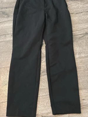 Svarta raka kostymbyxor - Snygga svarta kostymbyxor med raka ben och bälteshällor. Byxorna har två passpoalfickor bak och är tillverkade i ett slätt tyg som ger en clean look. Perfekta för dig som vill ha en stilren och enkel byxa till garderoben.