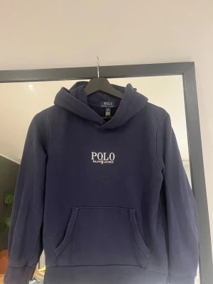 Mörkblå hoodie från Polo Ralph Lauren - Mörkblå hoodie från Polo Ralph Lauren med broderad vit logga på bröstet. Klassisk känguruficka framtill och huva. Skön och stilren modell i mjukt bomullsmaterial, perfekt för en avslappnad look. Original pris 1200