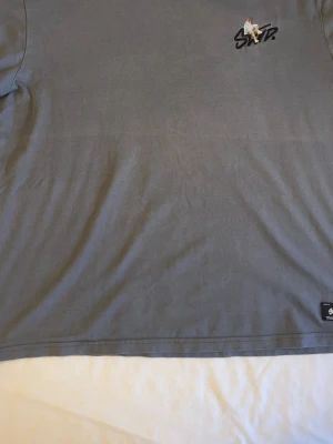 Grå t-shirt från SNTB med brodyr - Snygg grå t-shirt från SNTB med unik broderad detalj på bröstet som föreställer en sittande figur ovanpå loggan. Klassisk passform och mjukt bomullsmaterial. Perfekt för en chill och stilren look.