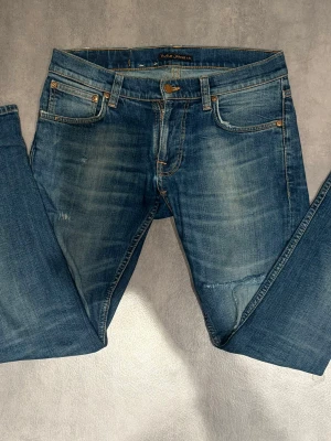 Nudie Jeans  - Snygga blå jeans från Nudie Jeans Co med klassisk femficksdesign och orangea sömmar. Jeansen har en lätt tvättad look och subtila slitningar framtill samt bakfickor med vågig brodyr. Perfekta för en avslappnad stil. Tight terry.