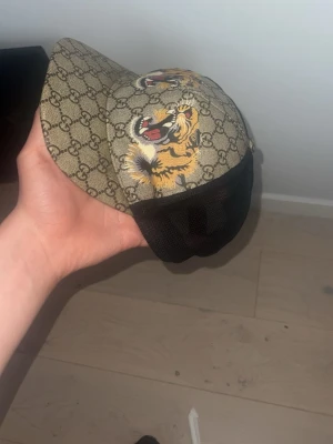 Gucci keps med tiger och GG-mönster - Säljer en snygg Gucci keps i beige med klassiskt GG-mönster och en cool tigerbrodyr framtill. Kepsen har svart mesh baktill och justerbart band med röd-gröna detaljer. Materialet är en mix av canvas och mesh, perfekt för en trendig streetwear-look.