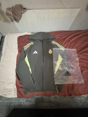 Real Madrid träningsjacka Adidas - Snygg grå träningsjacka från Adidas med Real Madrid-logga på bröstet. Jackan har huva, dragkedja och gula detaljer på ärmarna samt tre klassiska Adidas-ränder på axlarna. Perfekt för fotbollsträning eller chill. Materialet är troligtvis polyester.