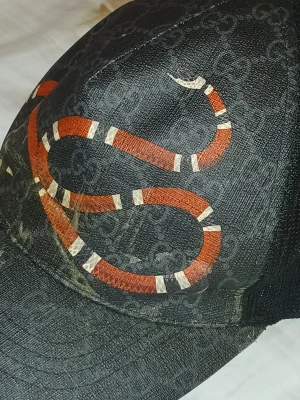 Svart Gucci keps med ormtryck - Cool svart keps från Gucci med ikoniskt GG-mönster och en röd/vit orm på framsidan. Baksidan har meshmaterial och justerbart band med blå och röda ränder. Perfekt för dig som vill sticka ut med en designeraccessoar.