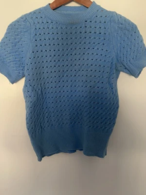 Ljusblå stickad topp med kort ärm - Snygg ljusblå stickad topp med korta ärmar och hålmönster över hela plagget. Tröjan har ribbstickade muddar vid ärmslut och nederkant samt rund halsringning. Perfekt för lager-på-lager och en chill look.