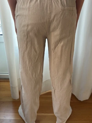 Beiga linnebyxor med resårmidja - Snygga beiga linnebyxor med avslappnad passform och resår i midjan samt dragsko. Byxorna har raka ben och en bakficka. Perfekta för en chill och stilren look. Materialet är luftigt linne som ger en skön känsla.