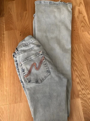 Ljusblå lågmidjade utsvängda (flared)Nelly jeans med slitna knän och detaljer där bak - Säljer ett par ljusblå jeans från Nelly, utsvängda ben och slitna knän. Bakfickorna har snygga rosa och vita broderade detaljer i form av vågor. Jeansen är gjorda i denim och har en cool sommar känsla