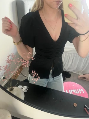 Svart v-ringad topp med snörning - Snygg svart topp med v-ringning och korta puffärmar. Toppen har snörning framtill som ger en cool, figurnära passform och en liten slits nedtill. Perfekt att styla med jeans eller kjol för en trendig look. Passar även en s