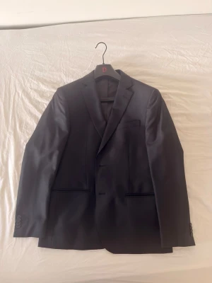 Svart kavaj från Dressmann - Stilren svart kavaj från Dressmann med klassisk passform. Kavajen har två knappar framtill, tre fickor och är helfodrad med slät insida. Perfekt till kostymbyxor eller jeans för en mer avslappnad look.