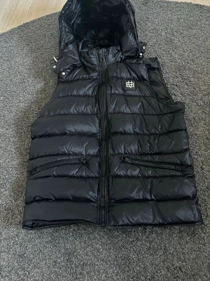 Svart dunväst med huva från bvaldi  - Snygg svart dunväst från bvaldi med avtagbar huva och två dragkedjefickor framtill. Västen har quiltad design och en broderad logga på bröstet. Obs västen har ett mega litet hål på insidan, jag personligen har aldrig tänkt på det och det är väldigt litet. Var inte räda att fråga om mer bilder eller något annat🤗 (pris kan gå ned vid snabb affär)