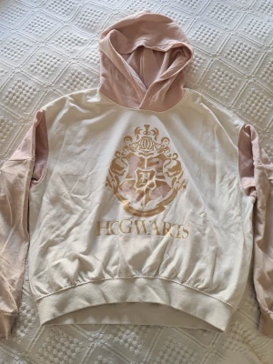 Hogwarts hoodie i beige och rosa - Säljer en snygg hoodie med Hogwarts-tryck i guld på bröstet. Hoodien är beige med ljusrosa huva och ärmar, och har ribbade muddar. Materialet känns mjukt och skönt. Texten har gått bort lite. Säljer då den är förlåten för mig och inte blivit använd på länge.