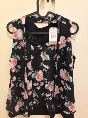 Blommig ärmlös blus  - Supersöt ärmlös blus  med svart botten och blommigt mönster i rosa, vitt och grönt. Blusen har v-ringning och ett lätt, luftigt material som passar perfekt till varmare dagar. Snygg att styla med jeans eller kjol.