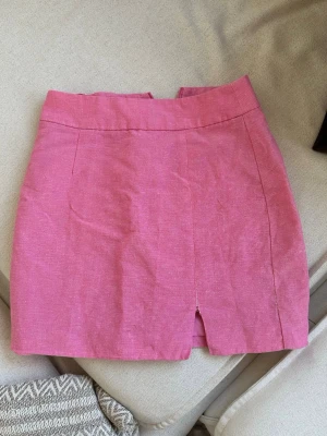 Rosa kjol med slits - Snygg rosa kjol i bomull med liten slits framtill som har spruckit lite (man kan säkert sy ihop lite lätt). Kjolen har en bred linning och diskreta sömmar för en stilren look. Perfekt att matcha med en enkel topp eller stickad tröja.