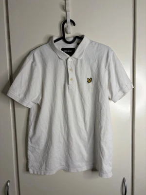 Vit pikétröja från Lyle & Scott - Klassisk vit pikétröja från Lyle & Scott med korta ärmar och gul logga på bröstet. Tröjan har krage och knäppning med tre knappar framtill. Perfekt för en stilren och avslappnad look.
