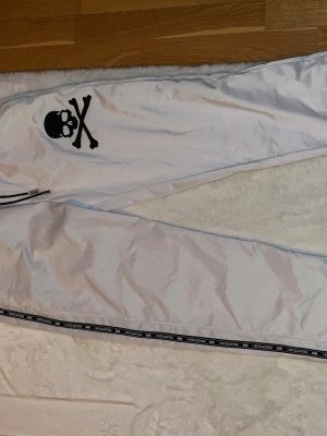 Vita mjukisbyxor från Philipp Plein - Snygga vita mjukisbyxor från Philipp Plein med svart dödskalletryck på benet och svarta detaljer längs bensluten. Byxorna har resår och dragsko i midjan samt dekorativa band med logga och årtal. OBS FINNS FLÄCKAR PÅ