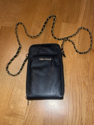 Svart axelväska  - Snygg svart axelväska i skinnimitation med guldfärgad kedjerem och logga framtill. Väskan har ett stilrent, avlångt format och dragkedja nertill. Perfekt för att bära det viktigaste med stil.