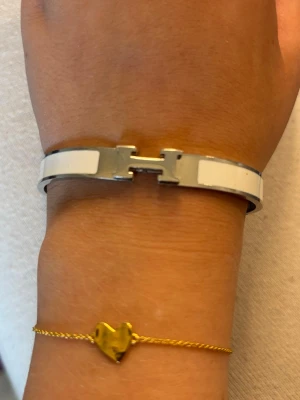 Hermès vit H-armband - Säljer ett stilrent armband från Hermès i vit och silverfärgad metall med den ikoniska H-detaljen framtill. Armbandet har en elegant och modern design som passar perfekt för dig som gillar exklusiva accessoarer.