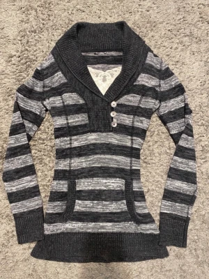 Randig stickad vintage Hollister tröja med stor krage - Mysig stickad tröja i svart och grått med breda ränder, stor sjalkrage och tre dekorativa knappar vid halsen. Tröjan har långa ärmar, ribbade muddar och en praktisk magficka framtill. Perfekt för dig som gillar en avslappnad men ändå snygg stil. den vita tröjan som är under tröjan kommer inte med!!!