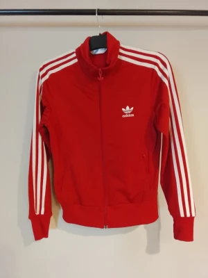 Adidas Originals track jacket NYSKICK - Röd track jacket från Adidas Originals med vita klassiska tre ränder längs ärmarna och vit logotyp på bröstet samt stort tryck på ryggen. Jackan har hög krage, dragkedja och ribbade muddar. Perfekt för sport eller streetwear. NYSKICK!