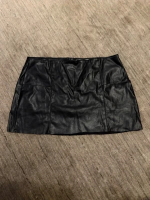 Svart kort kjol skinn - Snygg svart kort kjol i skinnimitation med stilren design. Kjolen har en rak passform och fickor fram. Storlek L men sitter mer som M.