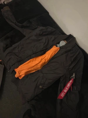 Svart bomberjacka med orange foder - Cool svart bomberjacka från Alpha Industries med klassiskt orange foder och ribbade muddar. Jackan har dragkedja framtill, fickor på sidorna och en ikonisk röd tag på ärmen. Perfekt för dig som vill ha en streetig och tidlös look.