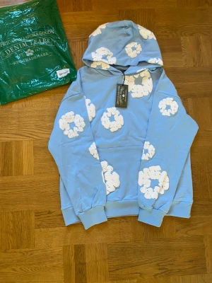 Blå hoodie med vita moln Denim Tears - Säljer en blå hoodie från Denim Tears med vita molnliknande tryck över hela tröjan. Hoodien har huva och ribbade muddar vid ärmslut och nederkant. Perfekt för dig som vill sticka ut med en unik och trendig look.