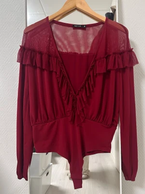 Vinröd meshbody med volanger - Vinröd body från My One med långa ärmar i mesh, djup V-ringning och volangdetaljer längs axlar och urringning. Tunt, stretchigt material som ger en feminin och trendig look. Perfekt för dig som vill ha något snyggt och bekvämt under kläderna.