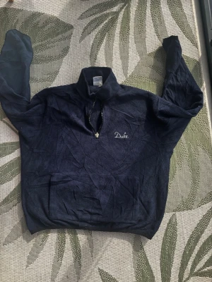 Mörkblå stickad tröja från Duke - Mörkblå stickad tröja från Duke med halv dragkedja och broderad logga på bröstet. Tröjan har hög krage och långa ärmar, perfekt för lager-på-lager-stil. Materialet känns mjukt och bekvämt.