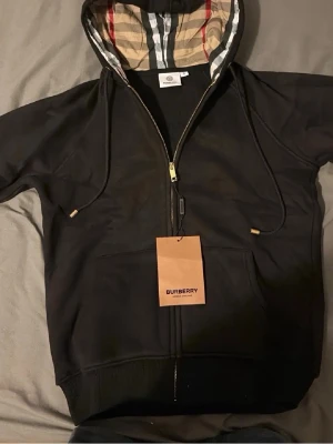 Svart Burberry hoodie med dragkedja - Svart hoodie från Burberry med klassiskt rutigt foder i huvan, dragkedja framtill och känguruficka. Snygga gulddetaljer på snörena och ribbade muddar. Perfekt för dig som vill ha en stilren och lyxig look. Sitter mer som S än M skulle jag säga 