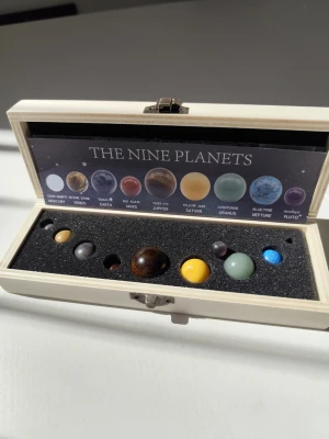 Planetset i träask – solsystemet - Snyggt set med nio planetkulor i olika färger och material, representerande solsystemets planeter. Kulorna ligger i en vit trälåda med svart skumgummi och medföljande plansch som visar planeternas namn och material. Perfekt som dekoration eller present till rymdintresserade.
